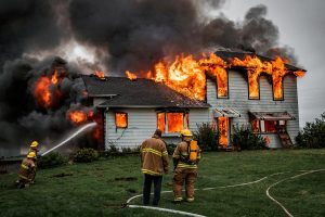A stick-frame white residential house engulfed in fire, with flames and smoke pouring out of the first and second story windows and roof as firefighters approach with water hoses