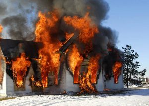 A one-story residential home engulfed in fire and billowing smoke on a winter day with a snowy yard